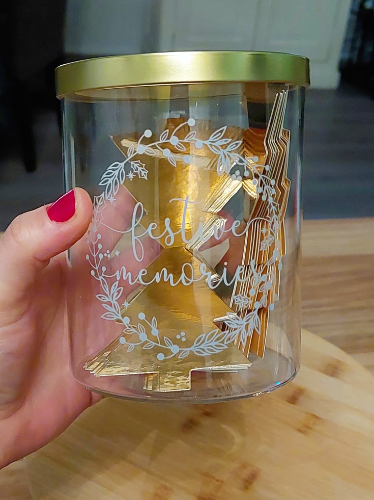Een hand met rode nagellak die een glazen pot met gouden deksel vast heeft met daarop gouden kerstboompjes voor Festive Memories. 
