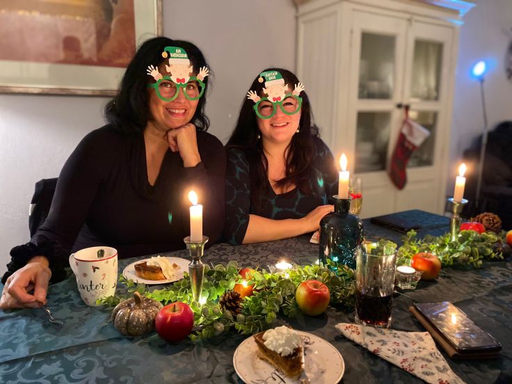 Twee vrouwen met kerstbrillen aan een feestelijk versierde eettafel met kaarslicht en kerstdecoratie.