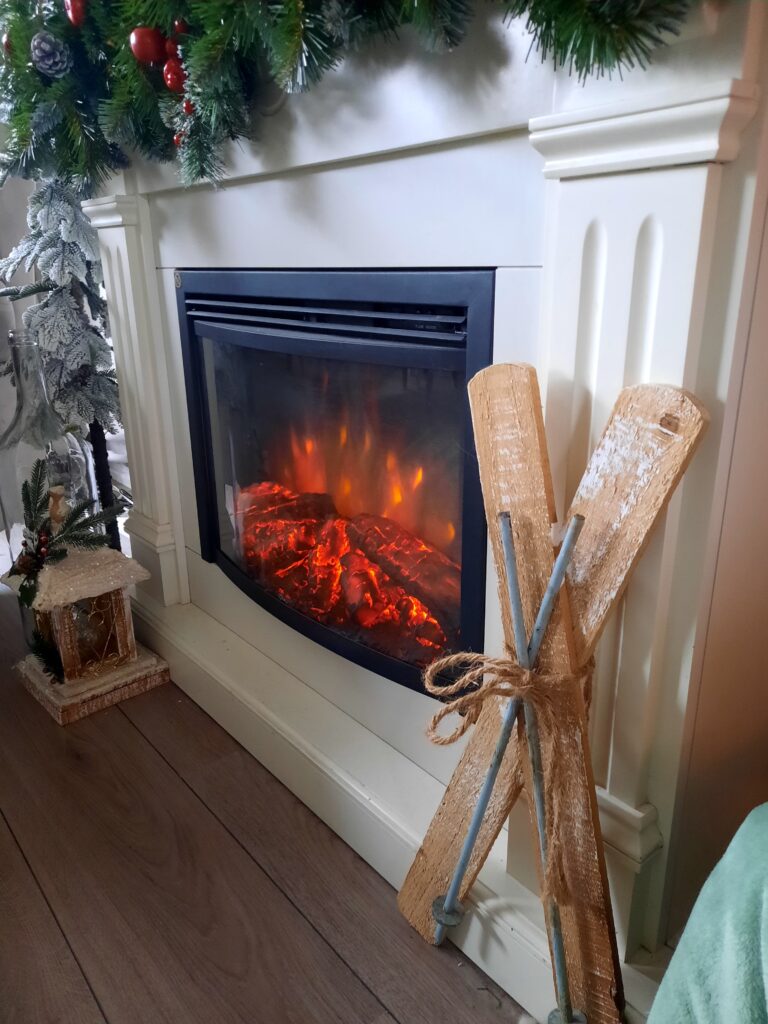 Een witte schouw met een brandende elektrische haard, versierd met groene kersttakken, rode kerstballen en houten mini-skietjes tegen de zijkant geleund.