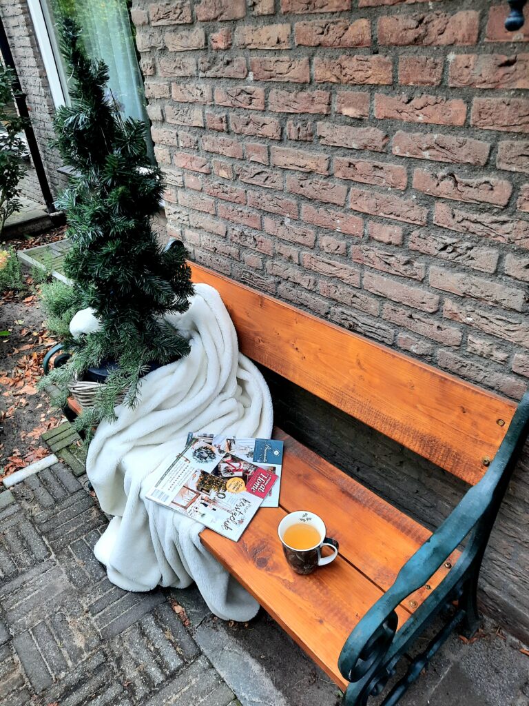 Houten bank, kopje thee, kersttijdschriften, plaid en kunstkerstboom in een groene en gele nazomer-setting.