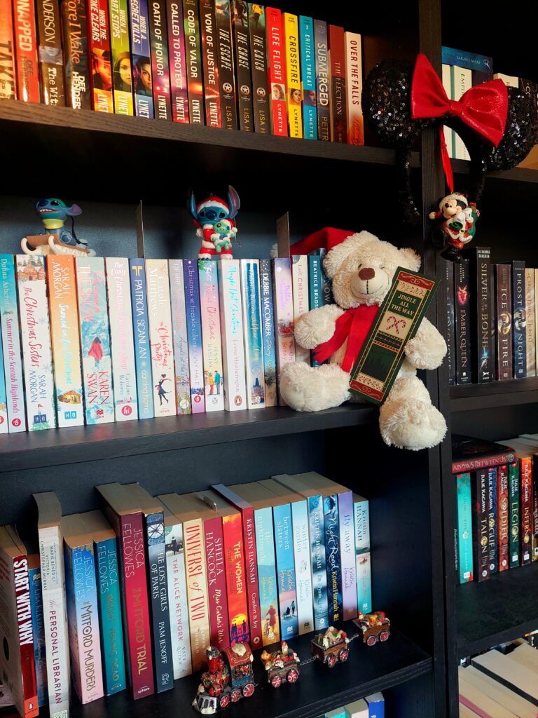 Boekenkast vol kleurrijke kerstromans met kerstdecoratie, waaronder een teddybeer met kerstboekenlegger, Disney Stitch-figuren en een miniatuur kersttrein.