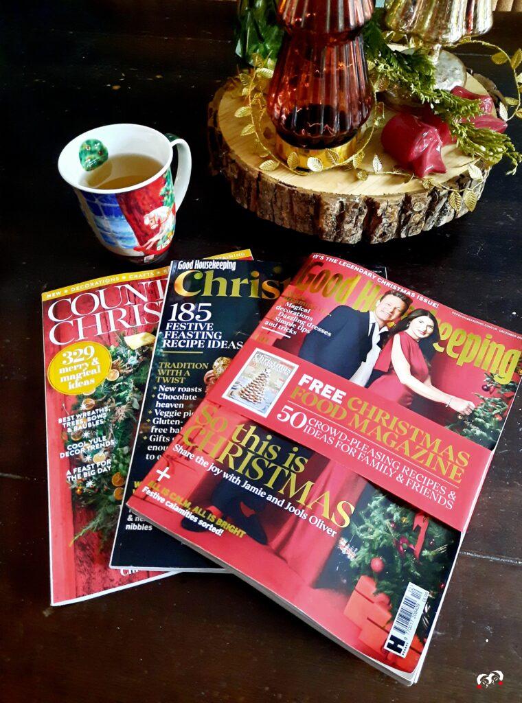 Tijdschriften met kerstinspiratie, waaronder Good Housekeeping Christmas en Country Christmas, op een donkere houten tafel met een kop thee en feestelijke decoratie. Perfecte sfeer voor het ontdekken van onze kerstthema’s 2025: Fabulous Burgundy & Christmas Classics.