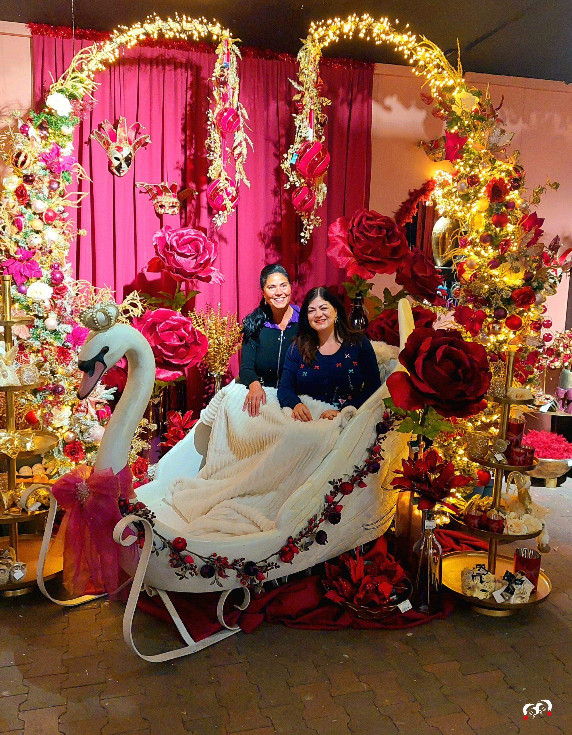 Carmen & Yardéna bij het luxe kerstdecor in La Vie en Rose-thema bij Coppelmans Oisterwijk met een witte zwaanslee, rode rozen, gouden accenten en fonkelende lichtjes.