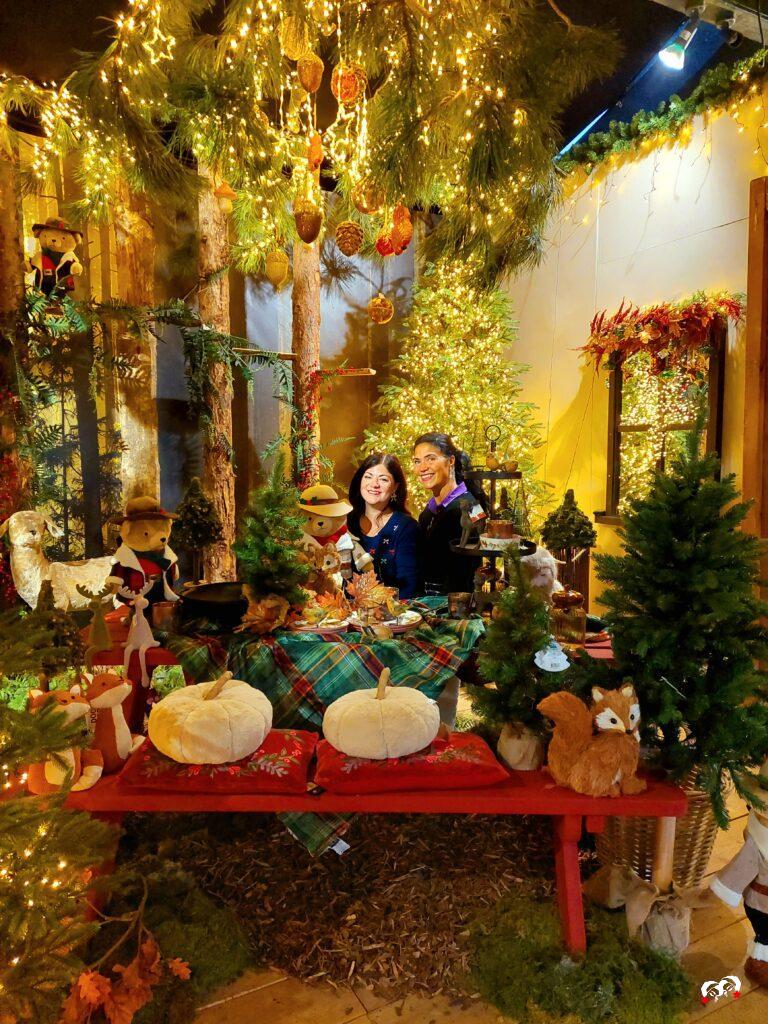 Carmen en Yardéna aan een feestelijk gedekte tafel vol herfst- en kerstdecoratie, omringd door fonkelende lichtjes, dennenbomen en bosdieren bij de kerstshow van Coppelmans Oisterwijk