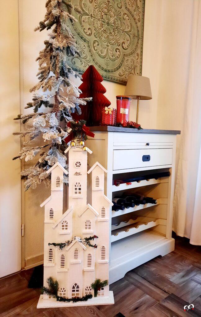 Landelijk wijnkastje met een wit houten huisje ervoor naast een besneeuwd kerstboompje en rode kerstdecoratie