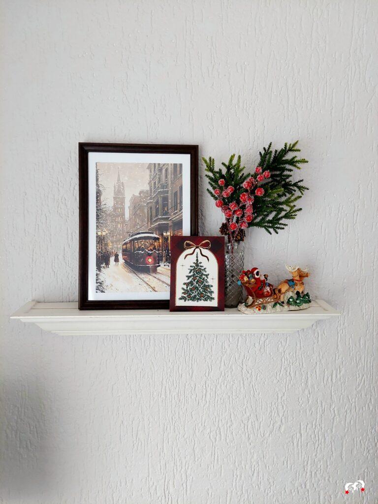 Wandplankje met kerstdecoratie en een poster in een lijstje. Dit is aangevuld met een kerstkaartje in tartan kleuren en wat extra kerstdecoratie