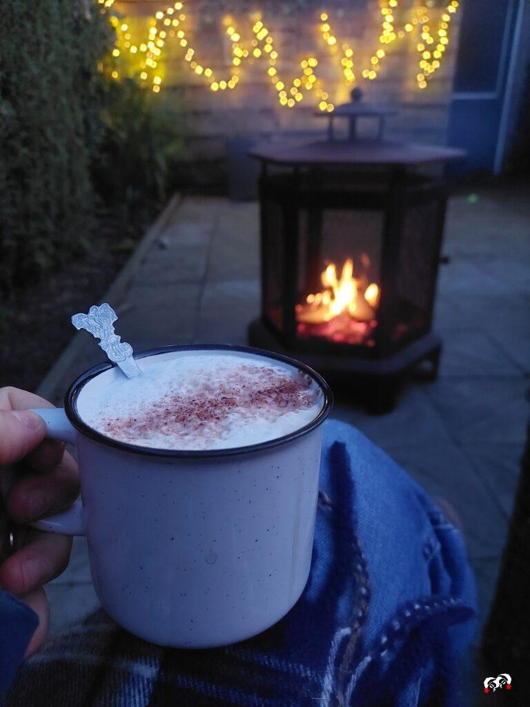 Koffiemok bij een vuurkorf in de avond, met tuinlichtjes, een omslagdoek en voeten in beeld.