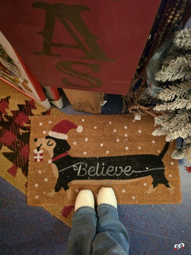 Kerstdeurmat met teckel met kerstmuts en de tekst “Believe”, met witte sneakers.