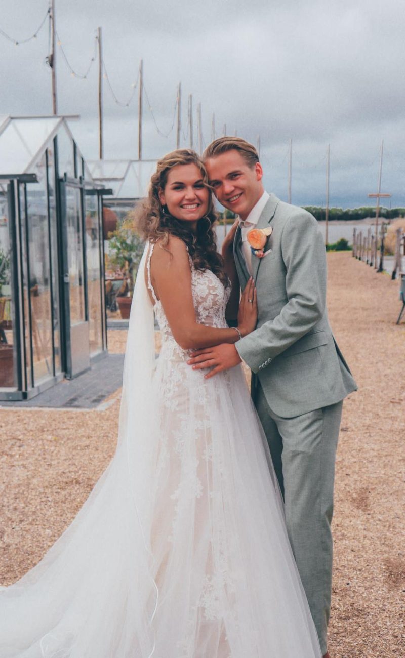 Bruidspaar poseert tussen kassen. Bruid draagt een mouwloze jurk met kant en een sluier. De bruidegom draagt een lichtgroen pak met een oranje bloem.