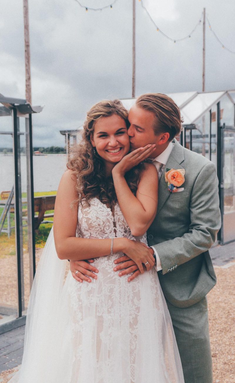 Bruidgom kust de bruid op haar wang en houd haar vast. Bruid draagt een mouwloze jurk met kant en een sluier. De bruidegom draagt een lichtgroen pak met een oranje bloem.