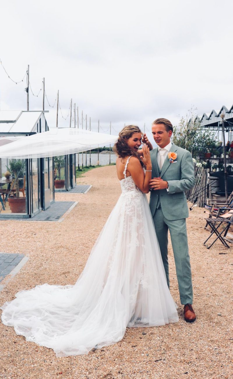 Bruidspaar poseert tussen kassen. Bruid draagt een mouwloze jurk met kant en een sluier. De bruidegom draagt een lichtgroen pak met een oranje bloem. Bruid houdt haar hand met ring omhoog.